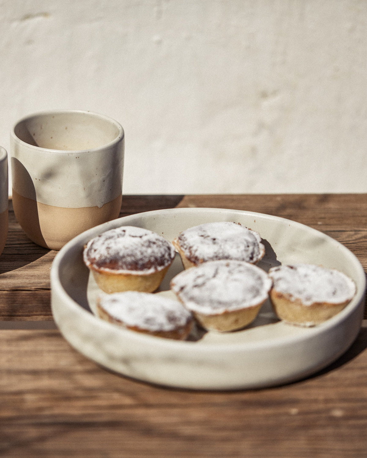 Cremefarbene Keramikschale mit vier mit Puderzucker bestäubten Mini-Törtchen auf einem Holztisch im Sonnenlicht, daneben ein zweitoniger Keramikbecher mit weißer Glasur und naturbelassenem Ton vor heller Wand.