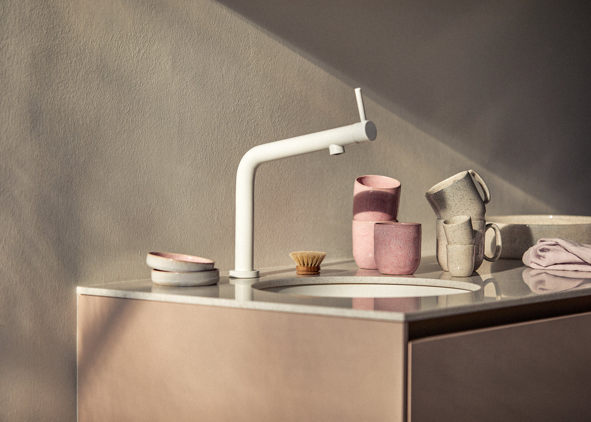 Modernes Küchenspülbecken aus hellem Stein mit weißer Design-Armatur, daneben rosa und cremegrau gesprenkeltes Keramikgeschirr (Becher mit und ohne Henkel sowie kleine Teller), eine Holzspülbürste und ein gefaltetes lavendelfarbenes Geschirrtuch im warmen Sonnenlicht.