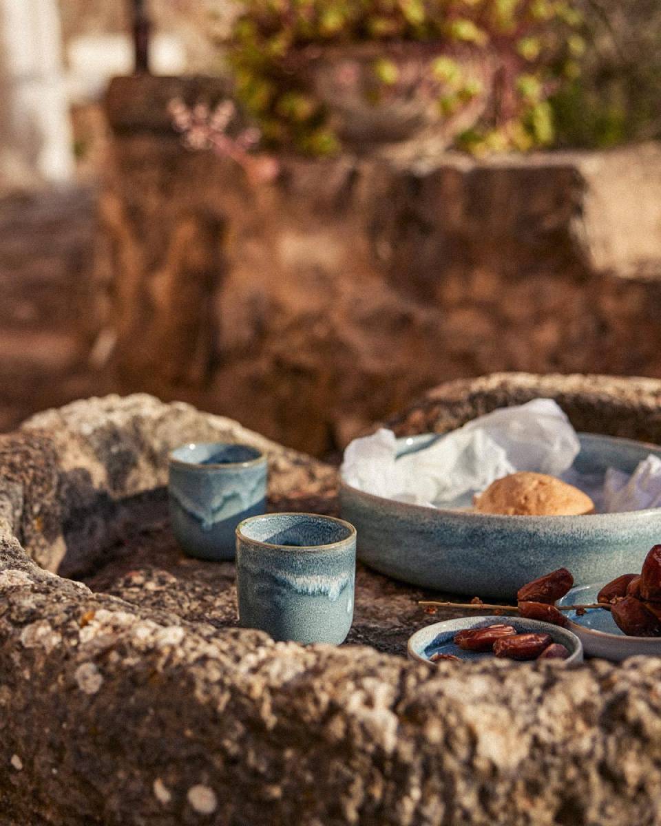 Zwei blaue Keramikbecher mit verlaufender Glasur und eine große blaue Servierschale mit einem Gebäckstück in Pergamentpapier auf einer rauen Natursteinmauer. Daneben ein kleiner blauer Teller mit getrockneten Datteln. Im Hintergrund warme Backsteinmauer mit grünen Pflanzen.