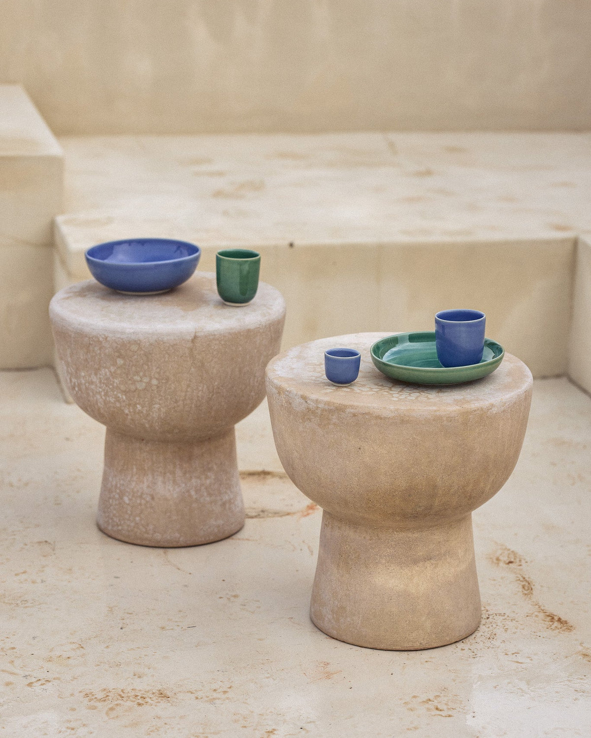 Zwei pilzförmige Beistelltische aus hellem Naturstein in einem mediterranen Außenbereich. Auf dem linken Tisch stehen eine blaue Keramikschüssel und ein grüner Keramikbecher, auf dem rechten ein grüner Teller mit einem blauen Becher und einem kleinen grünen Schälchen. Die leuchtenden Farben der glasierten Keramik setzen einen lebhaften Kontrast zur rauen, sandsteinfarbenen Oberfläche der Tische