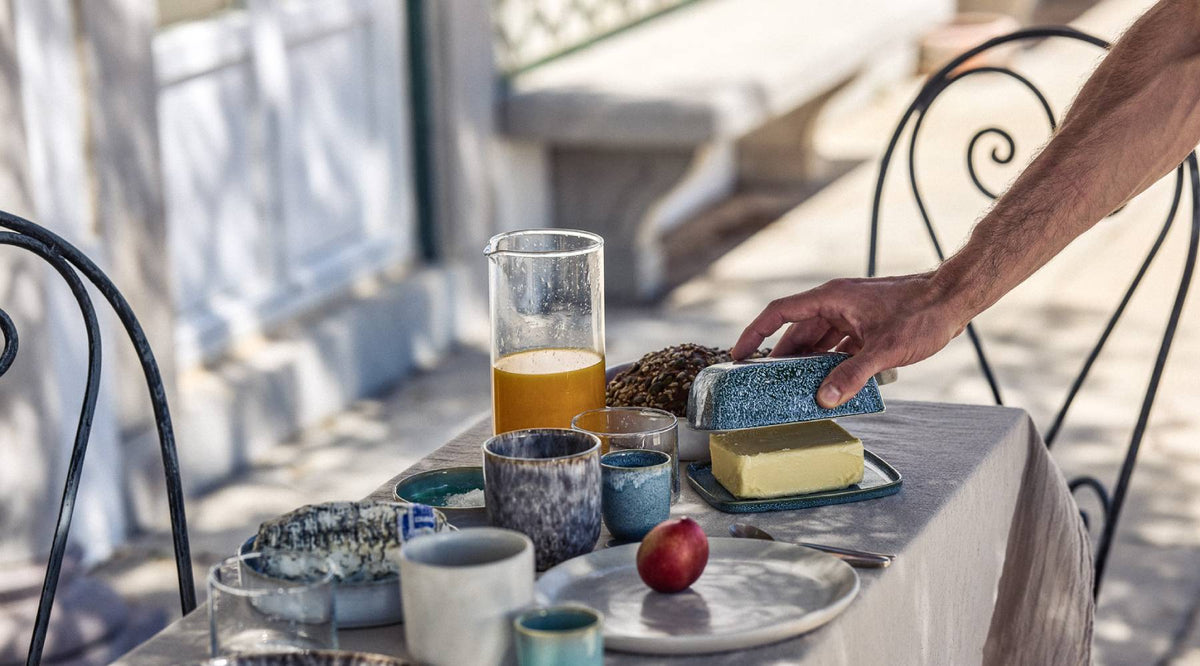 Hand greift nach einer blauen Keramik-Butterdose auf einem sonnigen Außentisch mit grauem Leinentischtuch. Auf dem Tisch stehen verschiedene blaue und graue Keramikbecher und -schalen, ein Glaskrug mit Orangensaft, eine Nektarine auf einem Teller sowie ein Käsestück. Beidseitig schwarze schmiedeeiserne Stühle, im Hintergrund eine helle Hausfassade.
