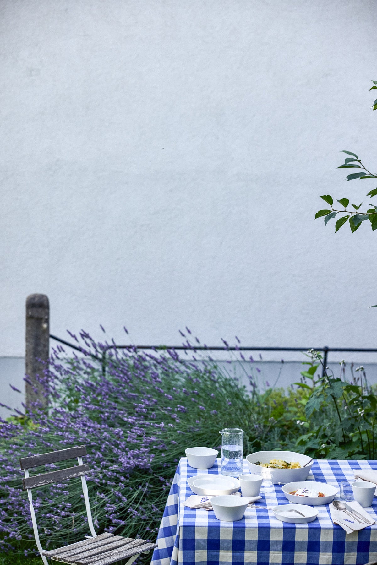 Gedeckter Gartentisch mit blau-weiß kariertem Tischtuch, darauf weiße Keramikschalen mit Speisen, ein Glaskrug mit Wasser und Besteck. Im Hintergrund blühender Lavendel vor einem schwarzen Metallzaun und einer weißen Hauswand, rechts ein Holzklappstuhl.
