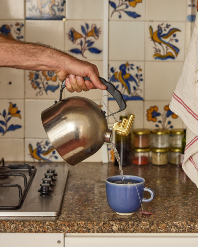 Hand gießt heißes Wasser aus einem silbernen Edelstahlkessel mit Messingausguss in eine blaue Keramiktasse mit Teebeutel. Im Hintergrund eine Küche mit traditionellen blau-gelben Azulejo-Wandfliesen, Gewürzgläsern und einem Gasherd.