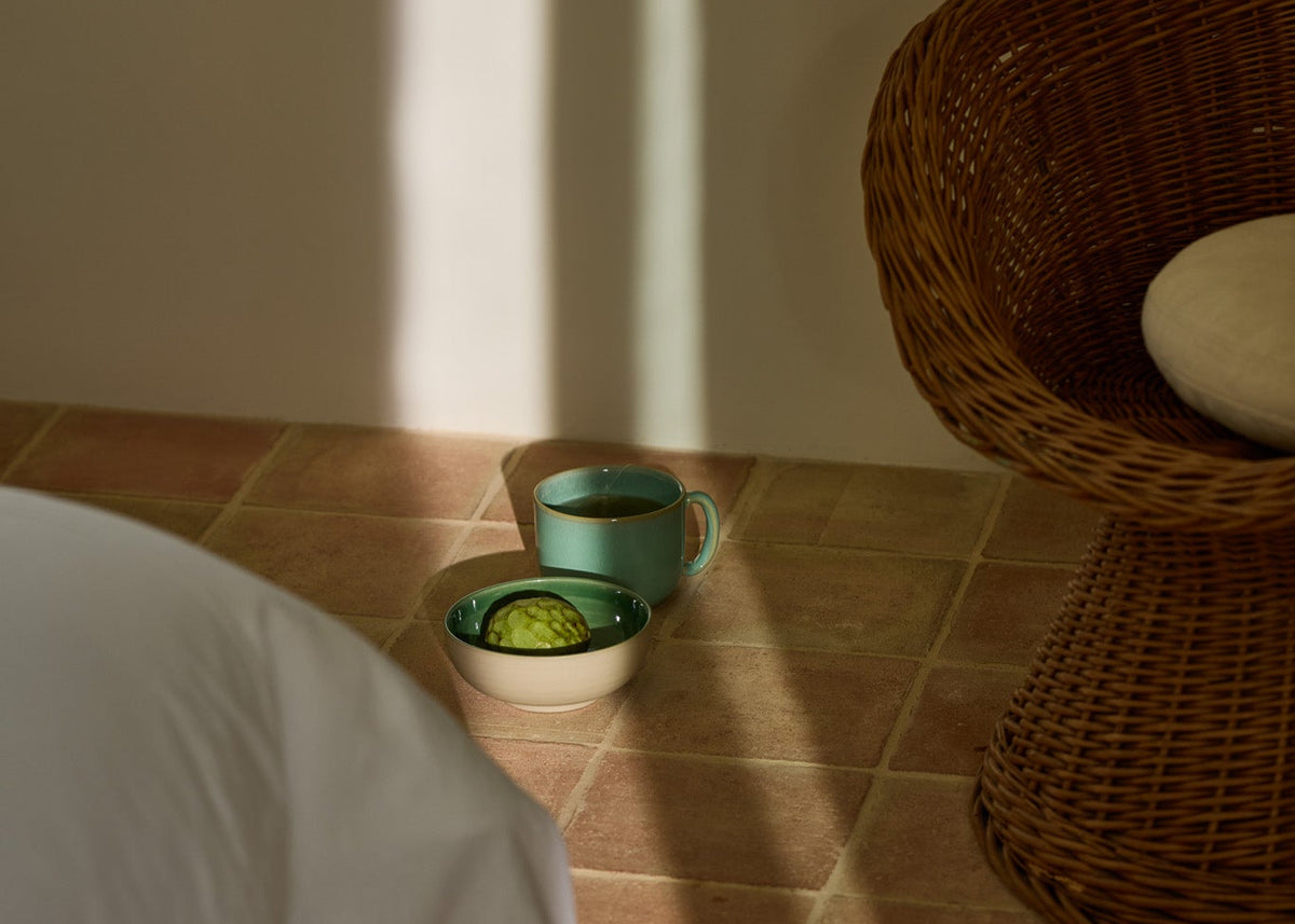 Türkisfarbene Keramiktasse mit Tee und eine cremefarbene Schale mit einer halben Avocado direkt auf einem Terrakottafliesenboden im warmen Sonnenlicht. Rechts ein Rattansessel, links ein weißes Sofa angeschnitten.
