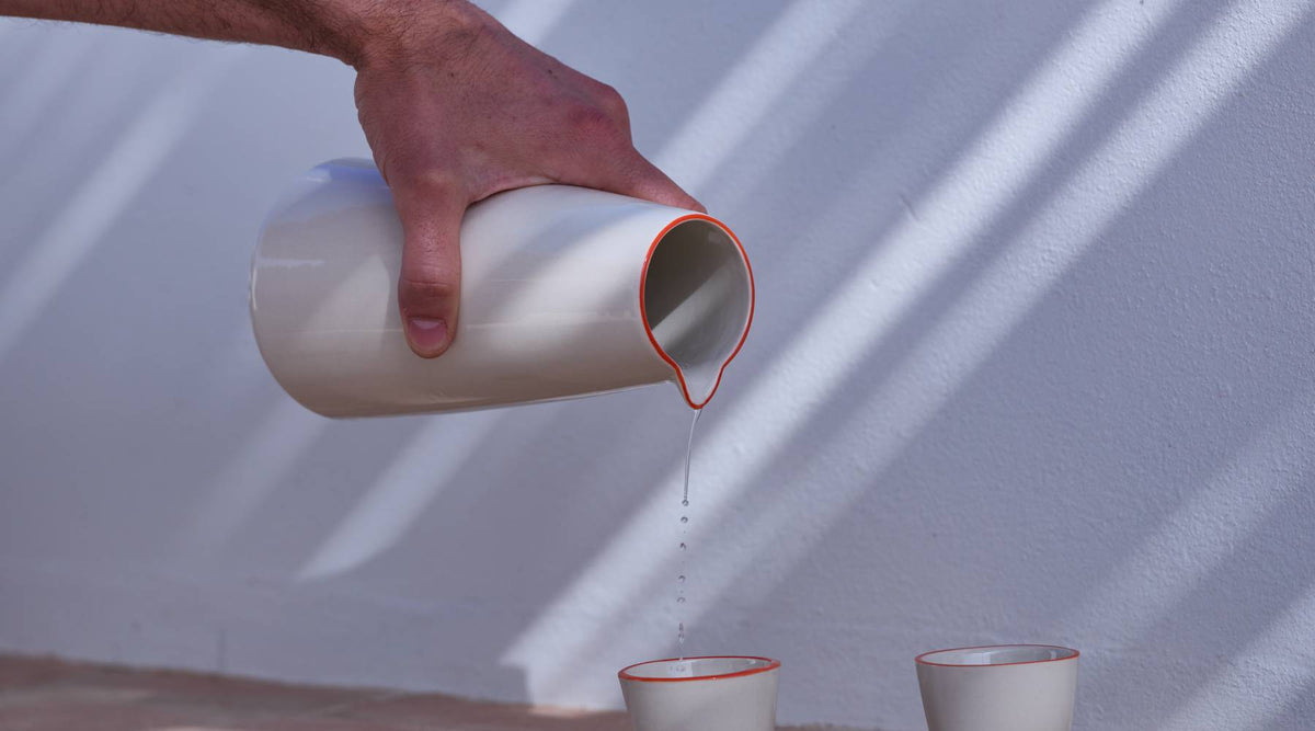 Hand gießt Wasser aus einem cremefarbenen Keramikkrug mit rotem Rand in einen passenden Becher darunter. Zweiter Becher daneben. Vor weißer Wand mit diagonalen Licht- und Schattenspielen.