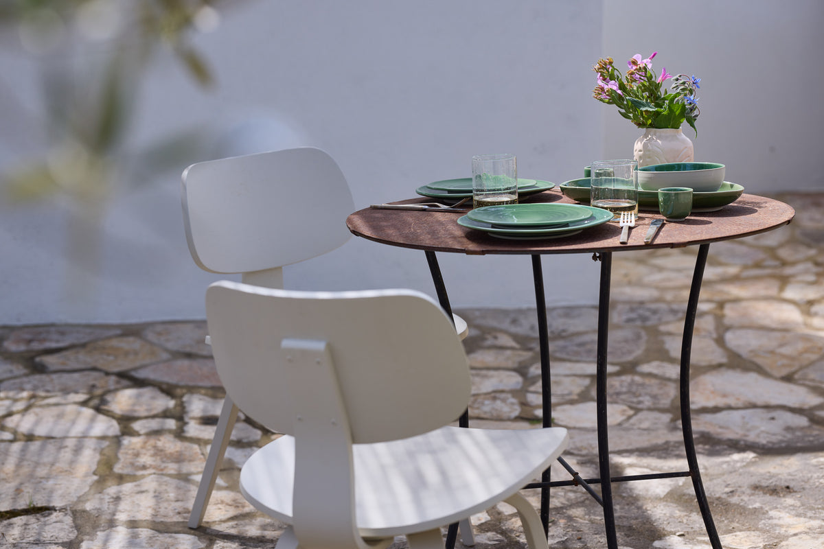 Kleiner runder Bistrotisch mit schwarzem Eisengestell auf einem mediterranen Steinpflaster-Außenbereich. Der Tisch ist für zwei Personen gedeckt mit grün-glasiertem Keramikgeschirr, gerippten Wassergläsern und dunklem Besteck. Eine rosafarbene Keramikvase mit bunten Wildblumen dient als Tischdekoration. Ein weißer moderner Holzstuhl steht vor dem Tisch, im Hintergrund eine weiße Wand im hellen Sonnenlicht.