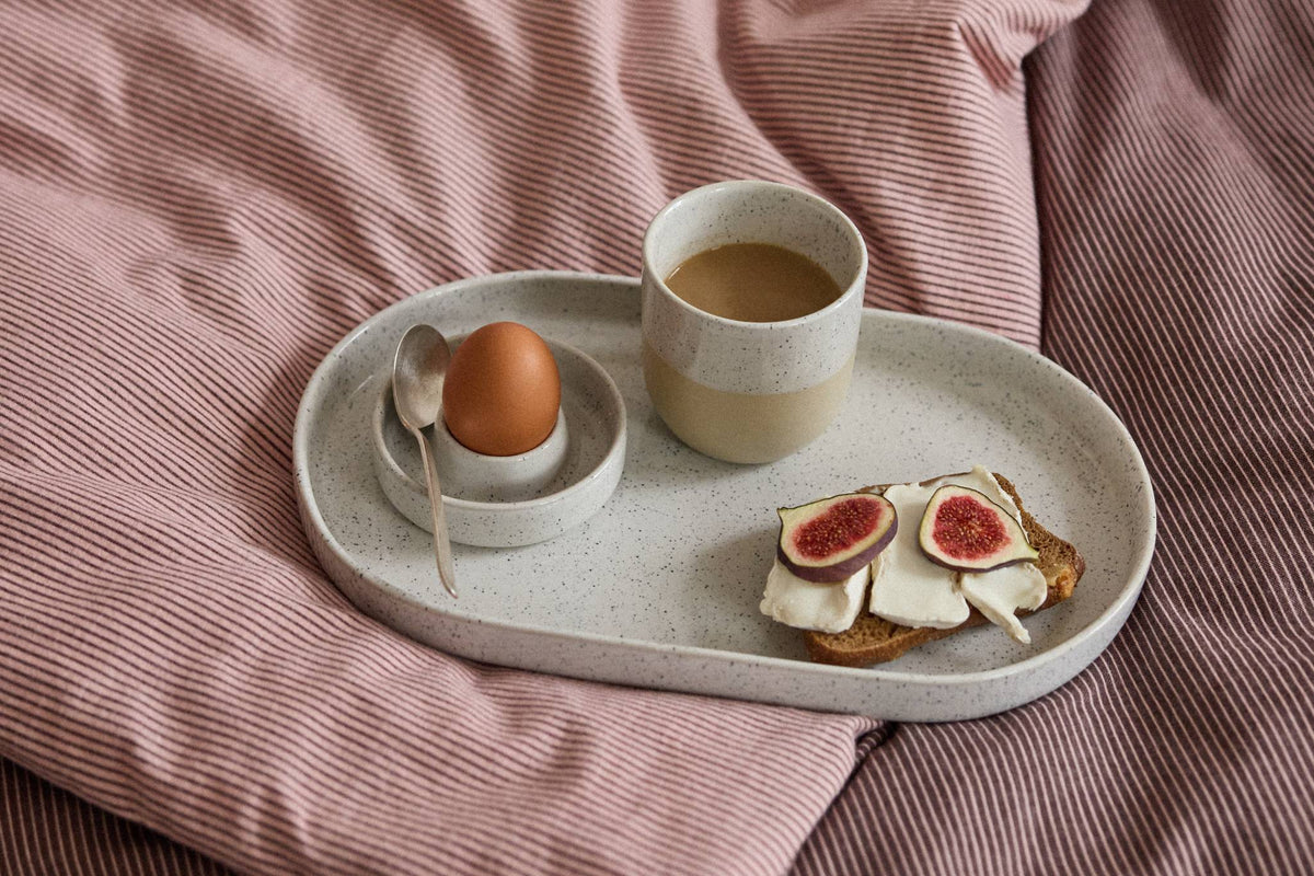Ovales, gesprenkeltes Keramiktablett mit Frühstücksei im Eierbecher, Becher mit Milchkaffee und Brot mit Feigen auf rosa Bettwäsche.