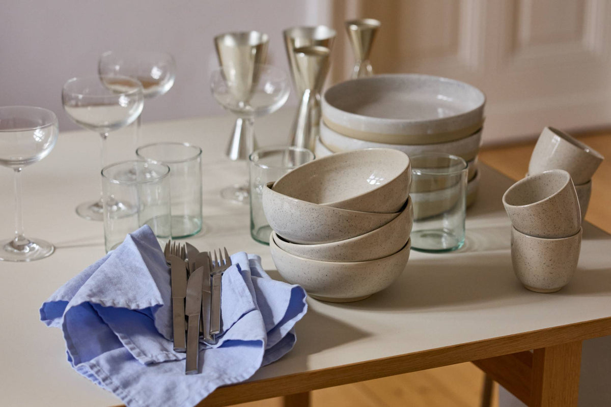 Holztisch mit heller Tischplatte in einem hellen Altbauzimmer mit Dielenboden und weißen Kassettentüren. Auf dem Tisch stehen gestapelte Keramikschalen, Teller und Becher in gesprenkeltem Beige sowie mehrere schlichte Wassergläser. Vorne liegt ein blau-weiß gestreiftes Leinentuch mit Besteck. Rechts steht ein dunkler Metallstuhl, warmes Sonnenlicht fällt in den Raum.