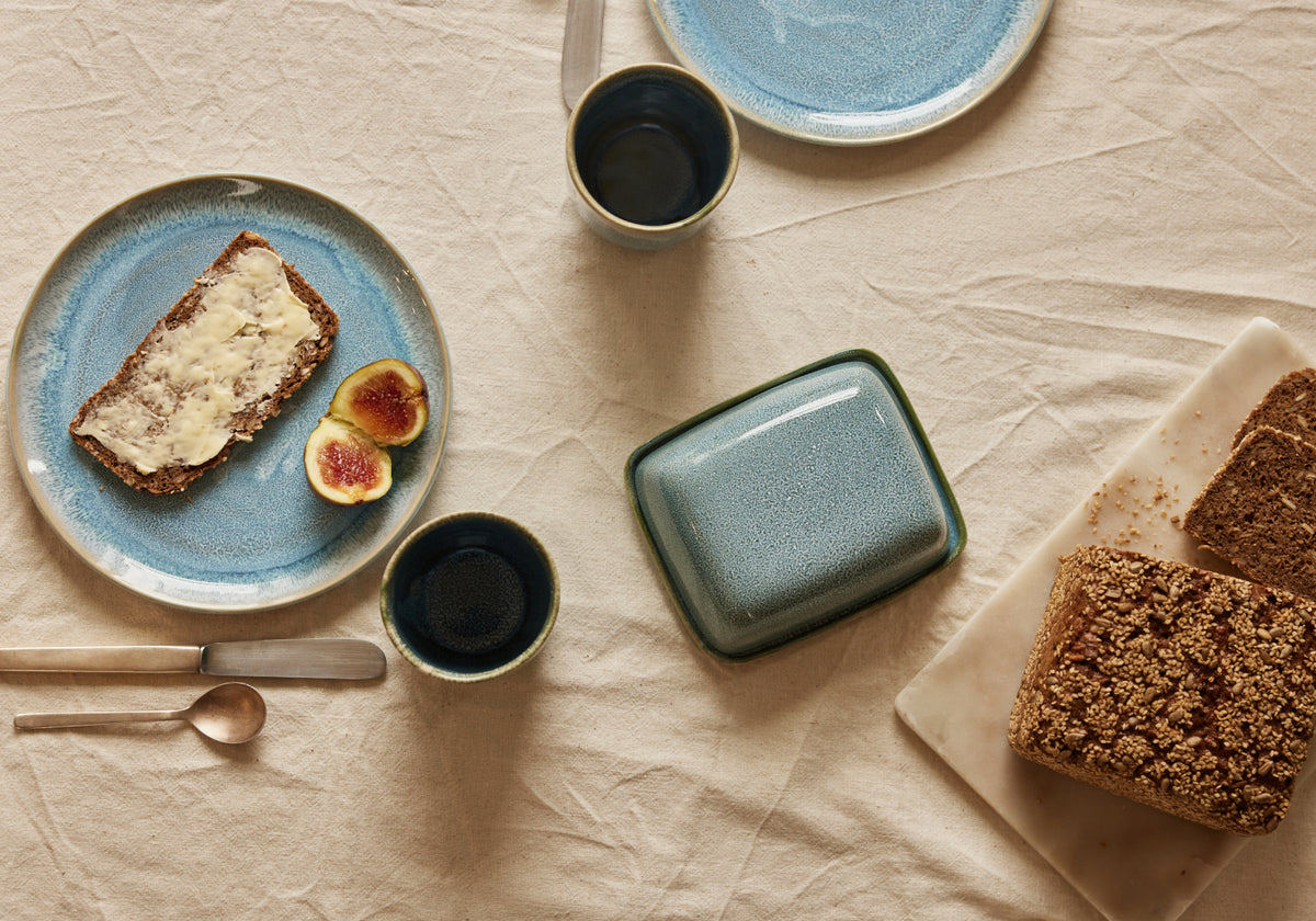 Vogelperspektive auf ein Frühstücksarrangement auf cremefarbenem Leinenstoff. Auf einem blau-glasierten Keramikteller liegt eine gebutterte Scheibe Körnerbrot mit zwei halbierten Feigen. Daneben eine türkis-grüne Keramik-Butterdose, zwei kleine dunkle Keramikbecher, ein weiterer blauer Teller im Hintergrund sowie aufgeschnittenes Körnerbrot auf einem weißen Marmorschneidebrett. Goldenes Besteck liegt links im Bild.