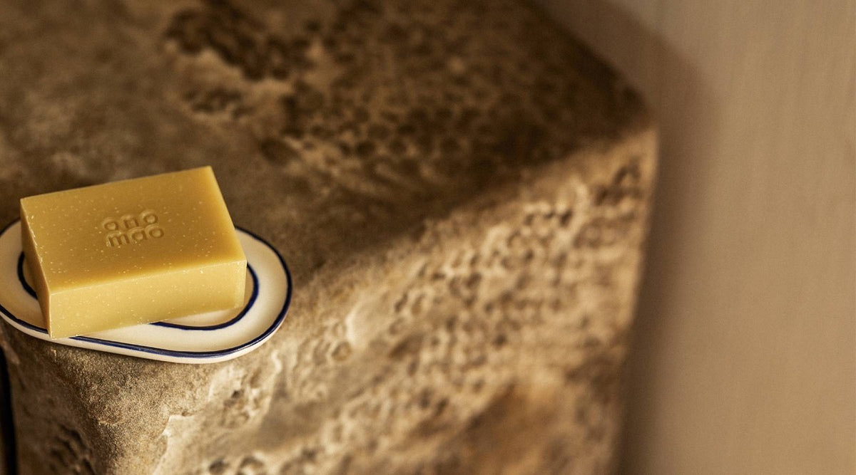 Rechteckiger, honiggelber Seifenblock mit geprägtem Schriftzug ‘ono mao’ liegt auf einer weiß glasierten, tropfenförmigen Keramik-Seifenschale mit dunkelblauem Rand; beides steht auf einer rauen, porösen Steinablage vor einer warm-beigen Wand.