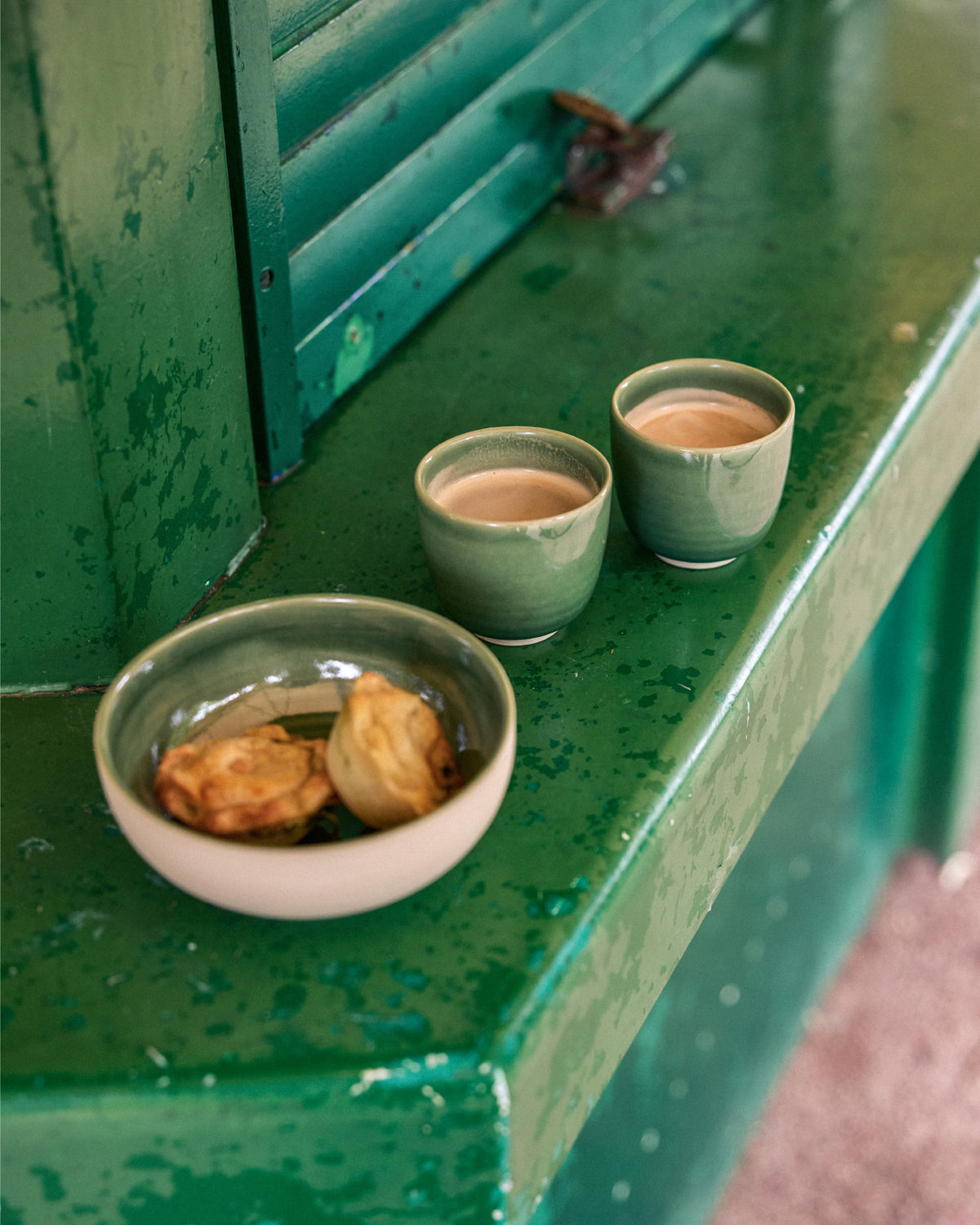 Zwei grüne Keramikbecher mit Kaffee und eine grüne Keramikschale mit kleinen Gebäckstücken auf einem verwitterten, grün lackierten Metallsims vor einem grünen Fensterladen.
