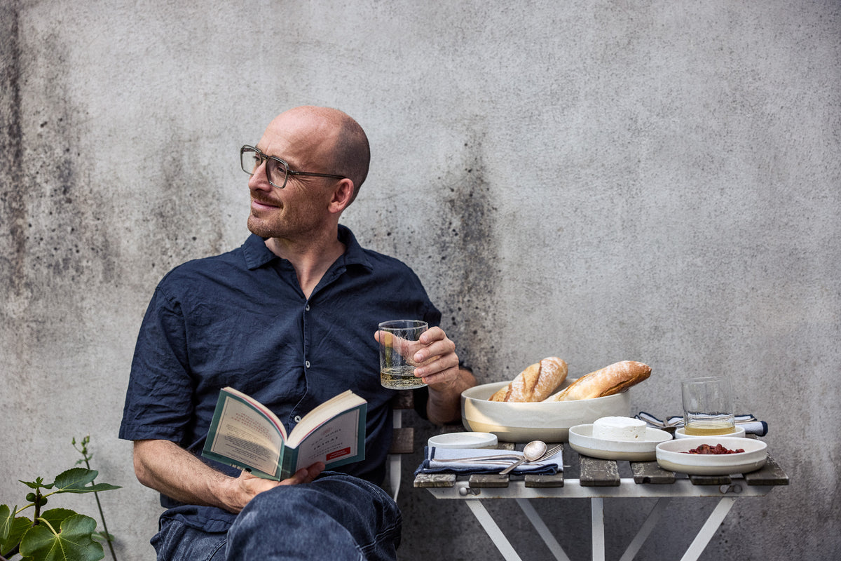 Mann mit Brille sitzt entspannt mit einem Buch und einem Glas Weißwein vor einer verwitterten Betonwand. Neben ihm ein Klapptisch mit cremefarbenem Keramikgeschirr, Baguettes, Frischkäse, getrockneten Tomaten und Besteck auf blau-weißen Stoffservietten.