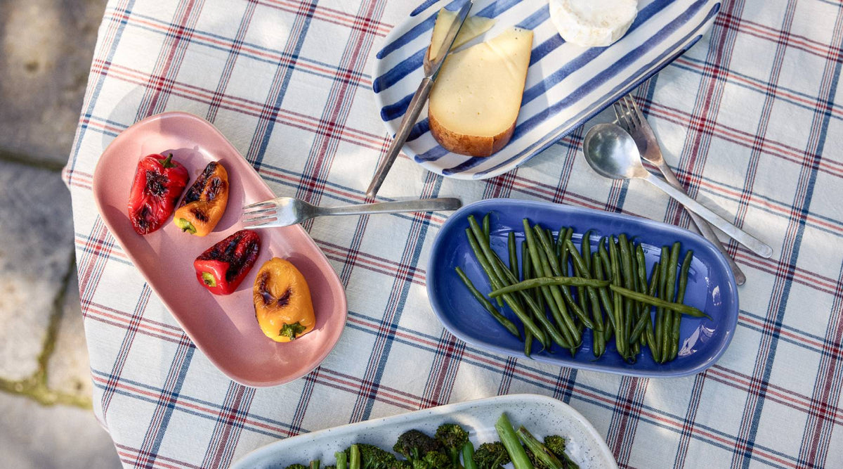 Vogelperspektive auf einen sommerlich gedeckten Tisch mit einem rot-weiß-blau karierten Tischtuch. Auf ovalen Serviertellern werden verschiedene Speisen präsentiert: gegrillte bunte Paprika auf einem rosafarbenen Teller, grüne Bohnen auf einem blauen Teller, gedünsteter Brokkoli auf einem weiß gesprenkelten Teller sowie überbackenes Brot auf einem blau-weiß gestreiften Teller