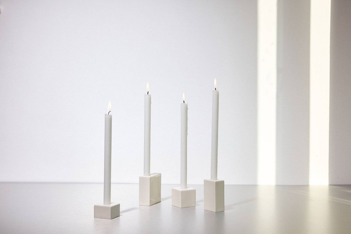 Drei brennende weiße Stabkerzen in minimalistischen quadratischen Keramikhaltern vor weißem Hintergrund