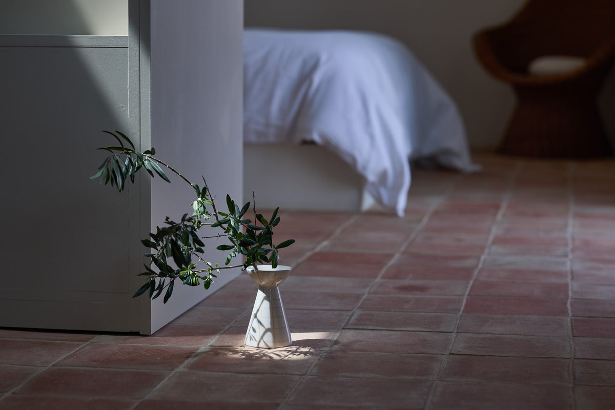 Eine schlanke, taillierte Keramikvase in Weiß mit einem Olivenzweig auf einem rustikalen Terrakottaboden. Ein Lichtstrahl fällt auf die Vase und wirft einen markanten Schatten. Im unscharfen Hintergrund ein Bett mit weißer Bettwäsche und ein Holzstuhl – eine ruhige, mediterrane Schlafzimmeratmosphäre.