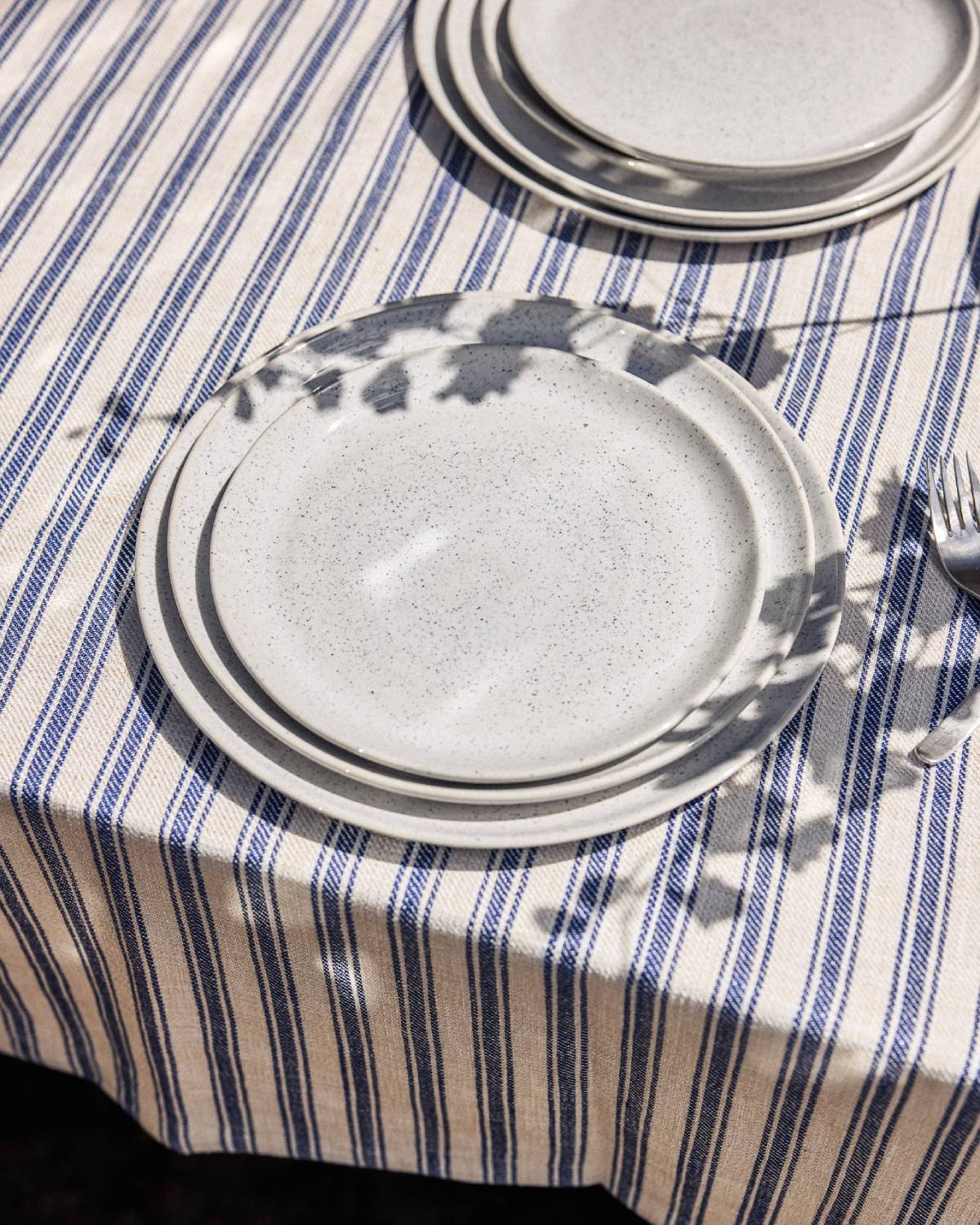 Gestapelte cremefarbene Keramikteller mit feiner Sprenkelung auf einem blau-weiß gestreiften Leinentischtuch im Sonnenlicht. Blattschatten fallen auf das Geschirr, rechts ist eine Gabel zu sehen.