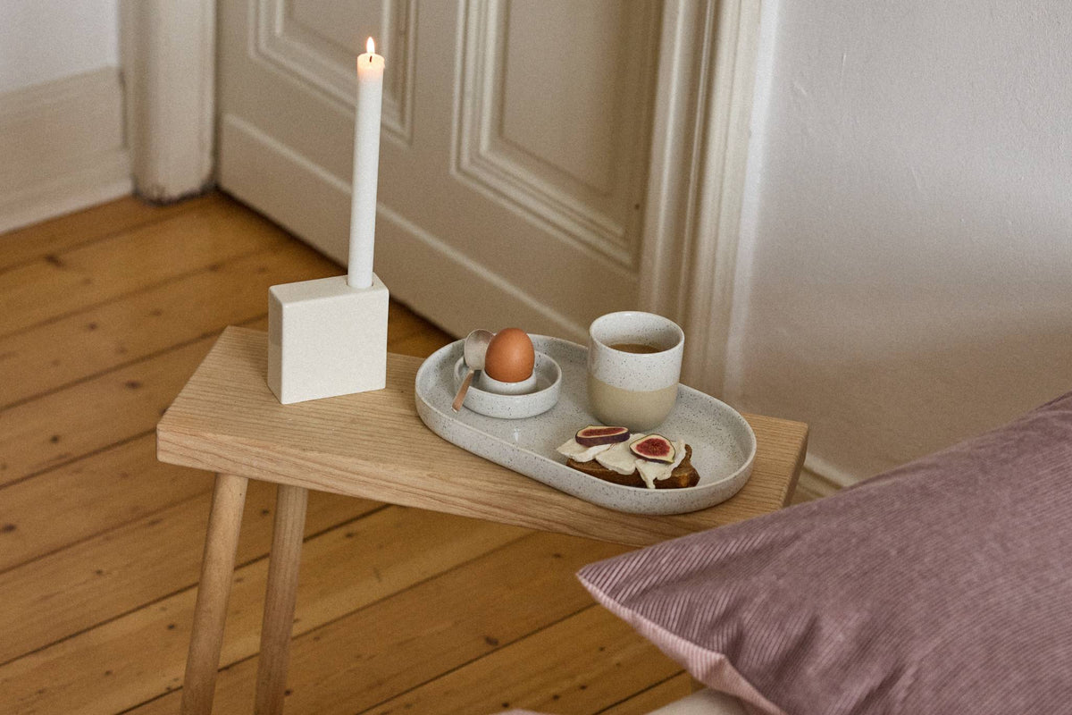 Ovales, gesprenkeltes Keramiktablett mit Ei im Eierbecher, Becher mit Kaffee und Feigenbrot auf einer Holzbank neben dem Bett.