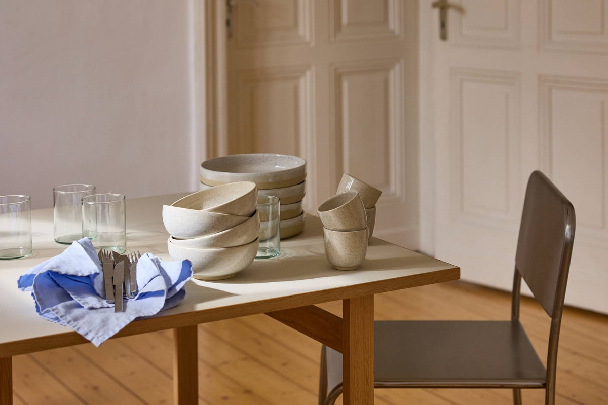 Holztisch mit heller Tischplatte in einem hellen Altbauzimmer mit Dielenboden und weißen Kassettentüren. Auf dem Tisch stehen gestapelte Keramikschalen, Teller und Becher in gesprenkeltem Beige sowie mehrere schlichte Wassergläser. Vorne liegt ein blau-weiß gestreiftes Leinentuch mit Besteck. Rechts steht ein dunkler Metallstuhl, warmes Sonnenlicht fällt in den Raum.