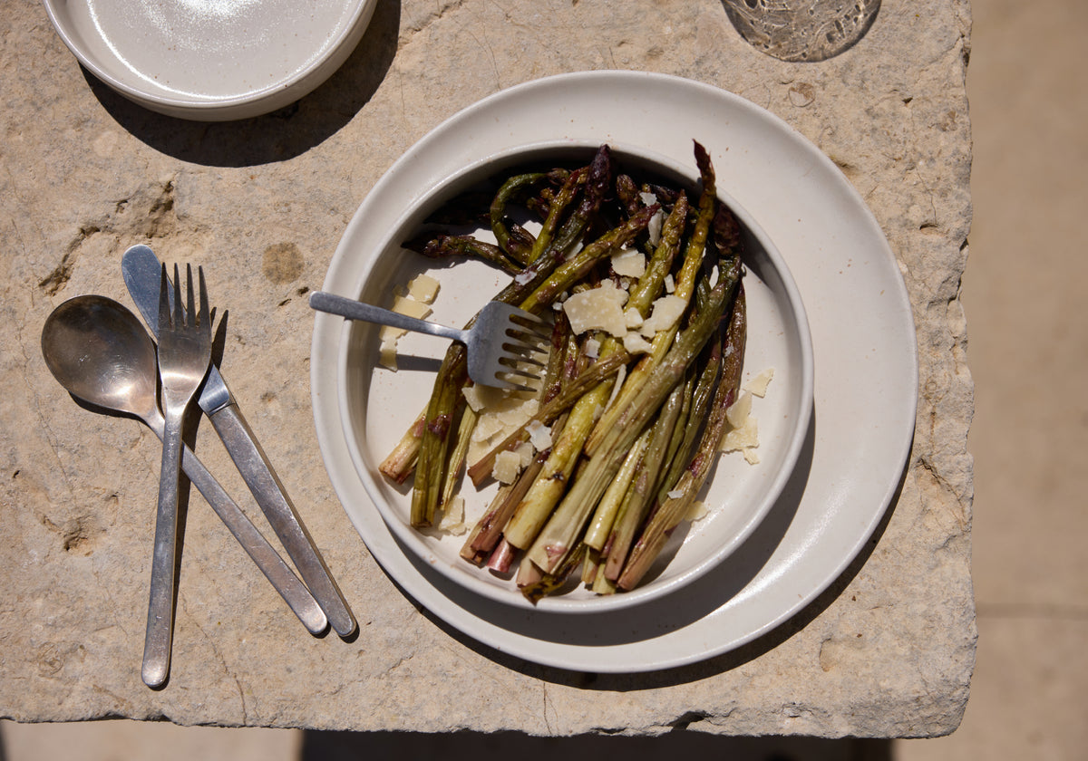 Gegrillter grüner Spargel mit Parmesanspänen auf einem cremefarbenen Keramikteller, serviert auf einer Natursteinplatte im Sonnenlicht. Daneben Gabel und Löffel aus Silber.