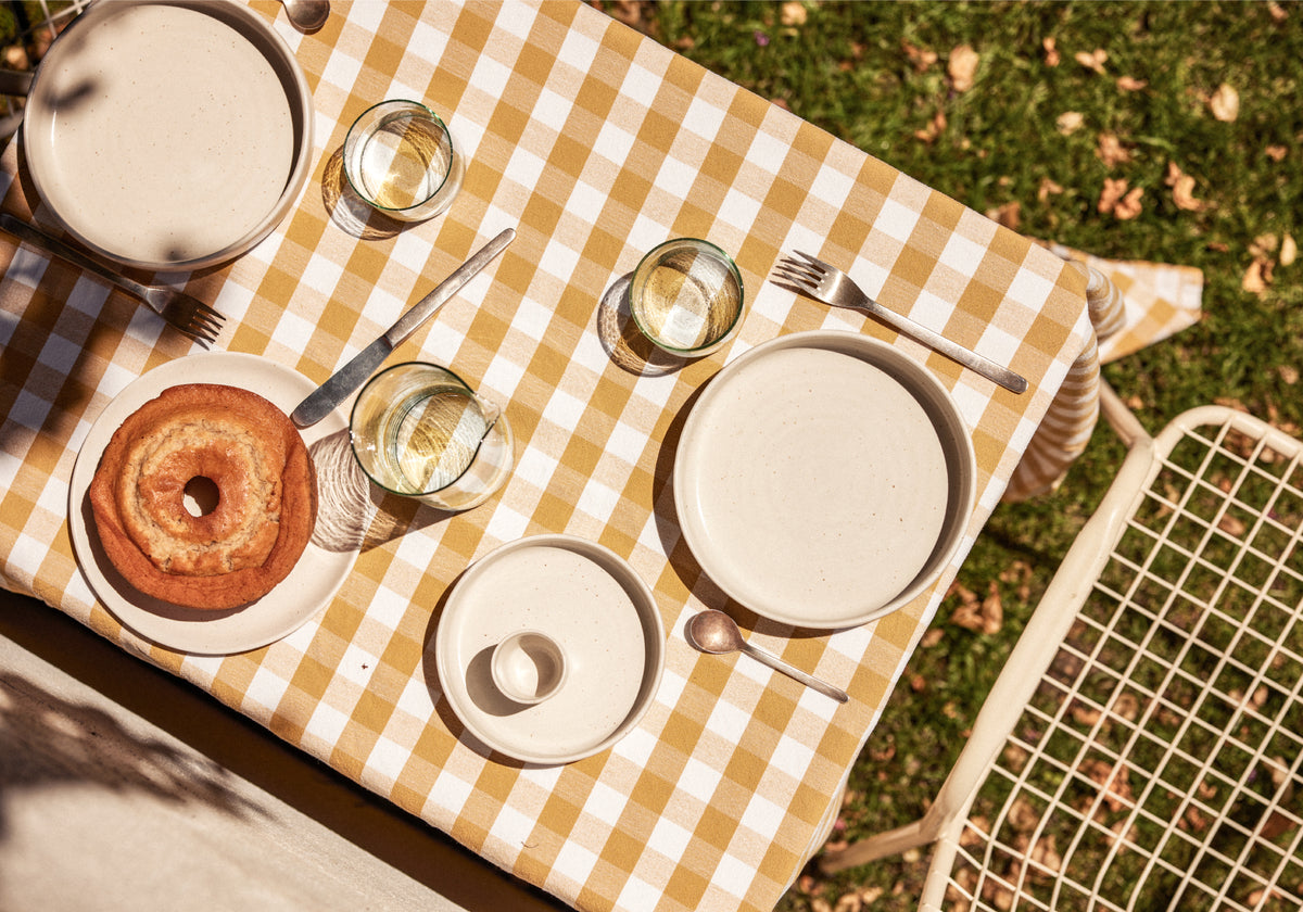 Gartentisch von oben mit ockergelb-weiß kariertem Tischtuch, darauf cremefarbenes Keramikgeschirr (Teller, Schalen und ein kleiner Zuckertopf), silbernes Besteck und drei Wassergläser. Links ein goldbrauner Gugelhupf auf einem Teller. Rechts ein weißer Metallgartenstuhl, im Hintergrund grüner Rasen mit Herbstlaub.
