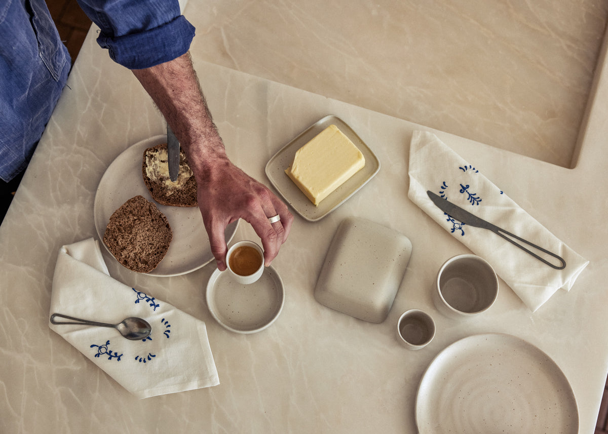 Vogelperspektive auf einen hellen Frühstückstisch. Eine Hand mit Ring greift nach einer kleinen Espressotasse auf Untertasse aus naturfarben gesprenkelter Keramik. Daneben ein Teller mit Körnerbrotscheiben, eine Butterdose mit frischer Butter, eine geschlossene quadratische Keramikdose sowie kleine Becher und ein Teller in gleichem Steinzeugton. Zwei weiße Leinenservietten mit blauer Stickerei und je einem Besteckteil vervollständigen das minimalistische Arrangement